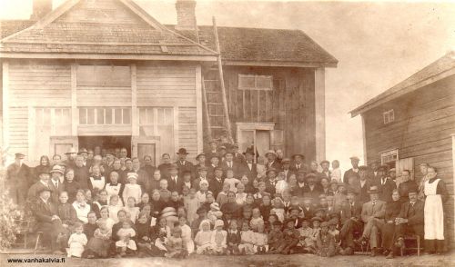 Hautajaiset Hillissä
Hautajaiset Salolla, Hillin kylällä. Salolta saadun tiedon mukaan kuva on talon vanhan isännän Antti Salon (s. 1844) hautajaisista kesällä 1918. 

Aatu Antinpoika Salo (s. 1880, Hilli 18) on kuvassa piharakennuksen oviaukossa seisomassa. Eturivin oikeassa reunassa istuu tuolilla Antti Antinpoika Salo (s. 1877, Hilli 18) ja vieressä vaimonsa Briita.  Briitan vieressä istuvasta hattupäisestä herrasta on useita nimiehdotuksia, mutta jos vertaa kuvaan ensimmäisestä kunnanvaltuustosta, niin hän on Juho Torppa. 

Toisessa rivissä on istumassa kaksi vaaleamekkoista tyttöstä. Oikeanpuoleinen on Laina Tikkanen vieressään äitinsä Edla (kuoli myöhemmin samana vuonna) ja hänen vieressään on Anna-Liisa Honkala. Hieman oikealle taaempana mustalakkinen, pienehkö mies on Juho Honkala vieressään poikansa Frans.

Takana äärimmäisenä vasemmalla aukinaisen oven vieressä seisoo tummahattuinen vanhempi mies, joka näyttää olevan Matti Hilli ja heti vieressään on vaimonsa Anna-Liisa Hilli (os. Peitso). Vasemman ovenpuoliskon kohdalla Fredrika ja Antti (punaparta) Hilli (Hilli 19) ja tuolilla istumassa vas. kolmas on tytär Lilja. Aivan vasemmalla edessä on Helli Rautomäki vieressään äitinsä Sofia (Satokoski 3) ja oikealle kolmen aikuisen jälkeen tyttönen on Martta Hilli (Hilli 22). Oviaukossa takana oikealla on Hanna Tuomisalo ja avonaisen ikkunen kohdalla aivan takana Roosa Renell (Ojakangas 3). Hänestä oikealle hattupäinen on Juho Ruokoja ja vähän alaspäin Lempi (Ruokoja 24). 

Rakennusten välissä ylärivissä mies pitelee valkopukuista lasta ja heti tämän lapsen alapuolella seisoo poika, jolla näyttää olevan valkoinen kauluspaita ja hattu päässä, hän on mahdollisesti Vilho Hilli, kuvaan jo tunnistetun Martta Hillin veli. Kuvassa ovat oletettavasti myös Vilhon ja Martan vanhemmat Gunda ja Jooseppi Hilli, mutta heitä ei vielä ole tunnistettu.

Keywords: Salo Hilli Torppa Tikkanen Honkala Rautomäki Tuomisalo Renell Ruokoja